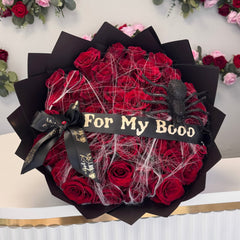 Bouquet of red roses with black ribbon and 'For My Boo' sign, surrounded by more flowers.
